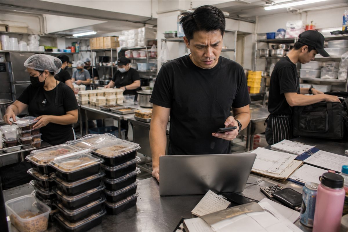 A caterer reviewing WhatsApp booking enquiries on a phone and laptop at a food preparation counter